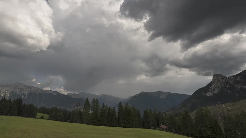 Dramatic storm clouds gather over a lush green meadow and pine forest in the majestic Dolomite mountains
