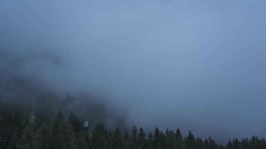 A dramatic timelapse shows thick, moody fog and clouds rolling through a mountain valley, obscuring the distant peaks and the forest below