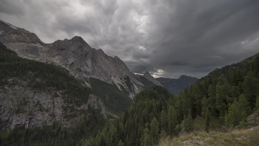 Dramatic storm clouds move across a majestic mountain range towering over a lush green forest valley