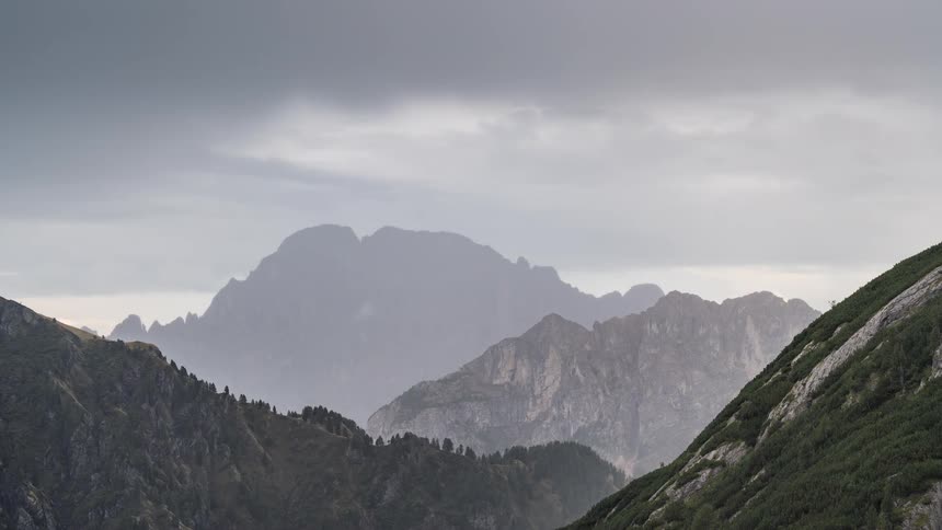 A majestic mountain range stands under a dark, cloudy sky as rain falls in the distance