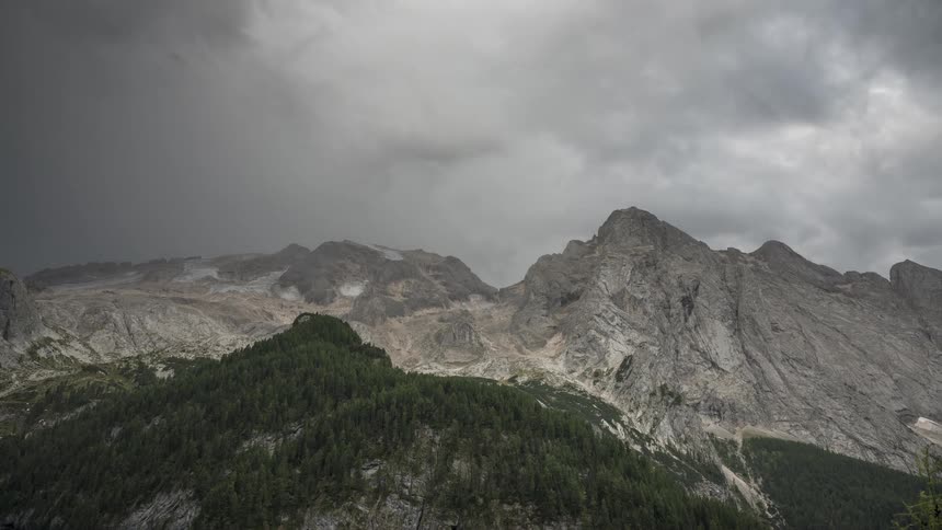 A dramatic timelapse captures dark, ominous storm clouds sweeping over a rugged mountain range with glaciers and pine forests