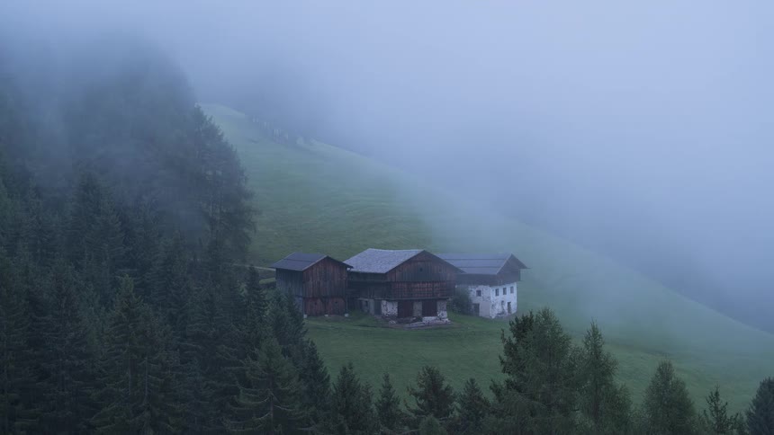 Thick fog rolls over a secluded, rustic farmhouse nestled on a green hillside surrounded by a pine forest
