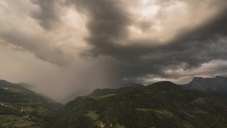 A dramatic timelapse captures a golden rainstorm moving through a lush green mountain valley under heavy, dark storm clouds