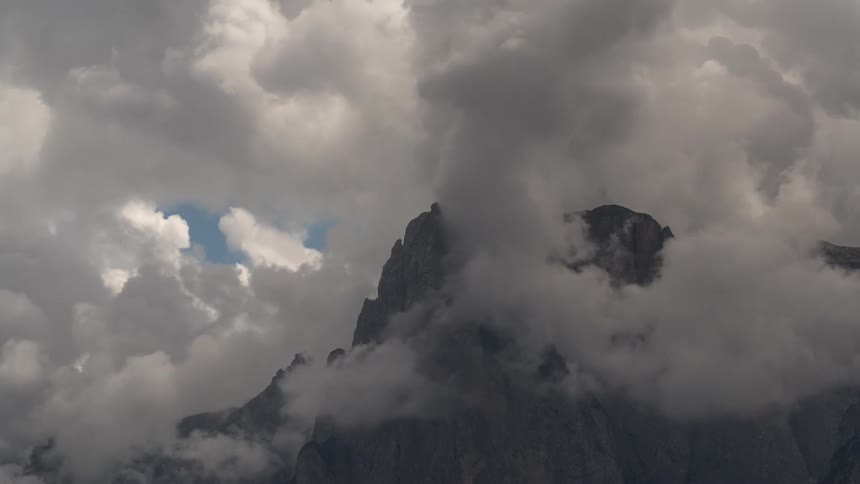 Thick, moody clouds slowly drift and swirl around a dramatic, rocky mountain peak
