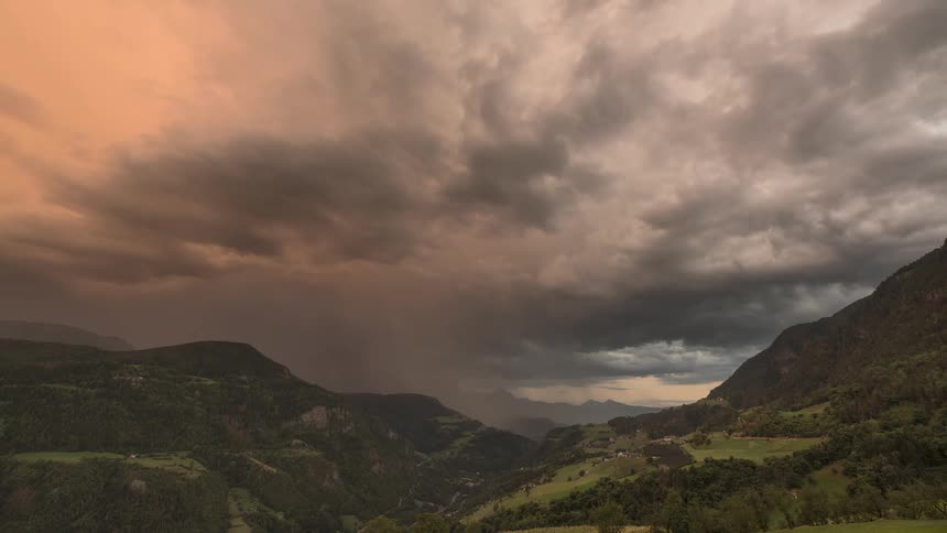 A dramatic time-lapse of a powerful thunderstorm with dark, churning clouds and rain moving through a lush mountain valley at sunset