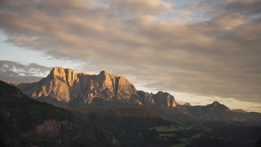 A beautiful day-to-night timelapse shows the sun setting behind a majestic mountain range, painting the clouds and peaks in golden and pink light
