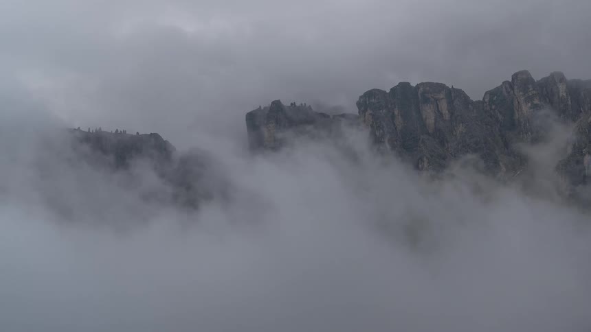 Timelapse of dense fog and clouds rolling through a rugged mountain range, gradually obscuring the rocky peaks