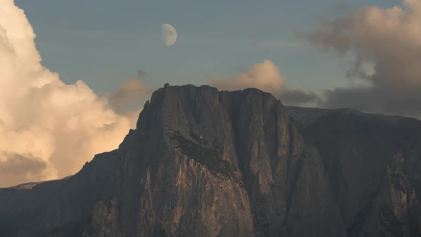 A majestic timelapse of a rugged mountain peak as golden hour clouds drift by and a half moon rises in the evening sky