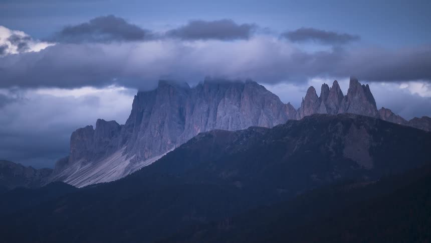 Dramatic clouds drift over the jagged peaks of the Dolomites mountain range at twilight