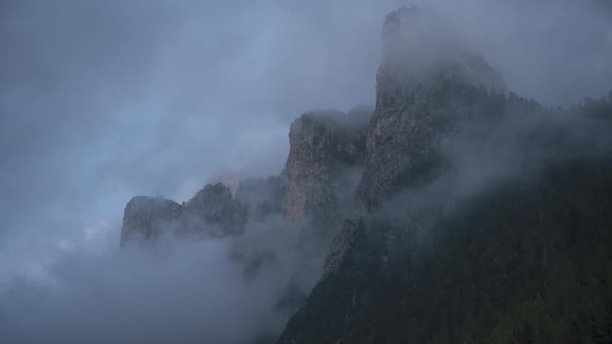 Moody clouds slowly drift past a rugged mountain peak, gradually revealing its majestic form at dusk