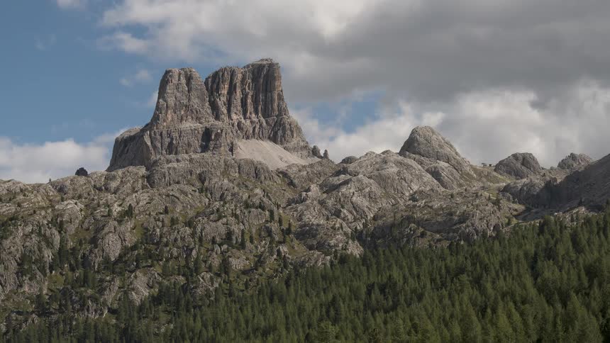 A static shot of a towering, rugged mountain peak above a dense pine forest, with soft clouds drifting across a blue sky