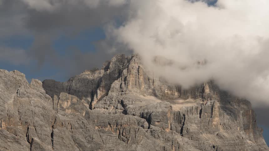 Wispy white clouds slowly drift over the rugged grey peaks of a mountain range on a sunny day
