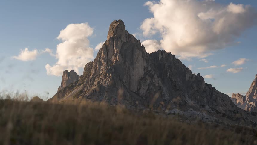 A stunning timelapse reveals clouds gracefully passing over a rugged mountain peak as golden hour light illuminates the swaying grass below