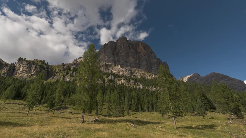 Clouds drift across a blue sky in a time-lapse over a towering rocky mountain and a green forest valley