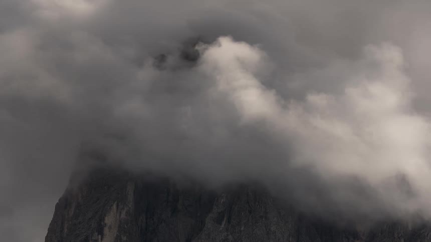 A time-lapse of thick, grey clouds swirling around a rugged mountain, slowly revealing its majestic peak