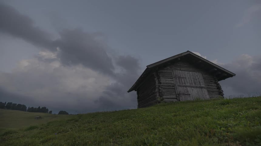A time-lapse captures dramatic clouds passing over a rustic log cabin on a grassy hill as daylight fades