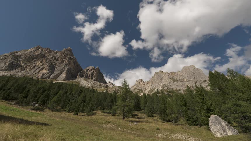 A scenic time-lapse of clouds moving across a deep blue sky over a majestic, rocky mountain range covered in green pine trees