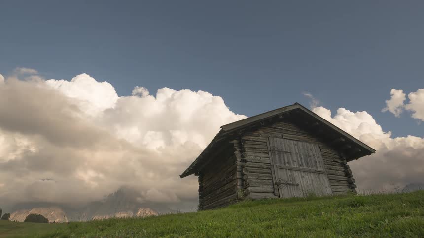 Dramatic clouds drift over a rustic log cabin perched on a lush green alpine meadow with mountains in the distance
