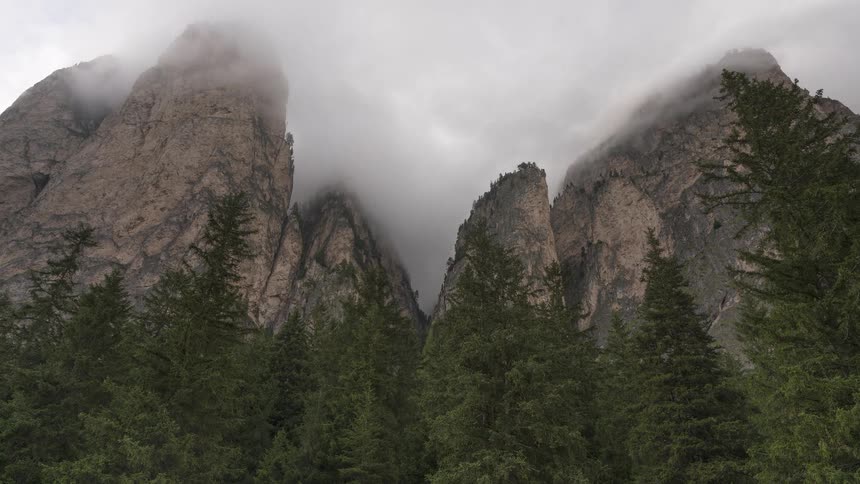 A dramatic time-lapse of thick clouds and fog rolling over jagged mountain peaks above a lush evergreen forest