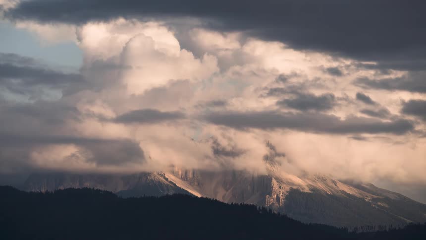 A dramatic time-lapse of thick, warm-hued clouds swirling around a majestic mountain peak at sunset