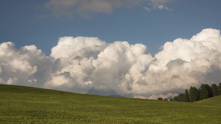 A majestic timelapse shows big, fluffy clouds rolling over a green alpine meadow with distant mountains