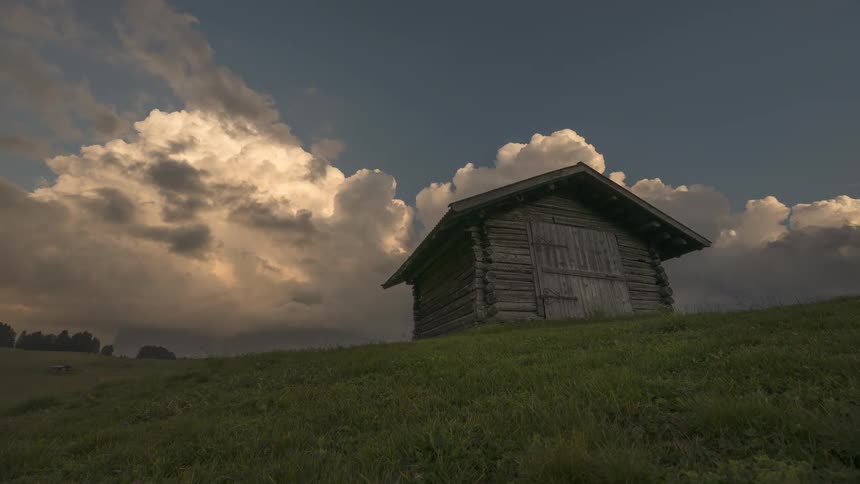A rustic wooden cabin sits alone on a grassy hillside under a dramatic, cloudy sky