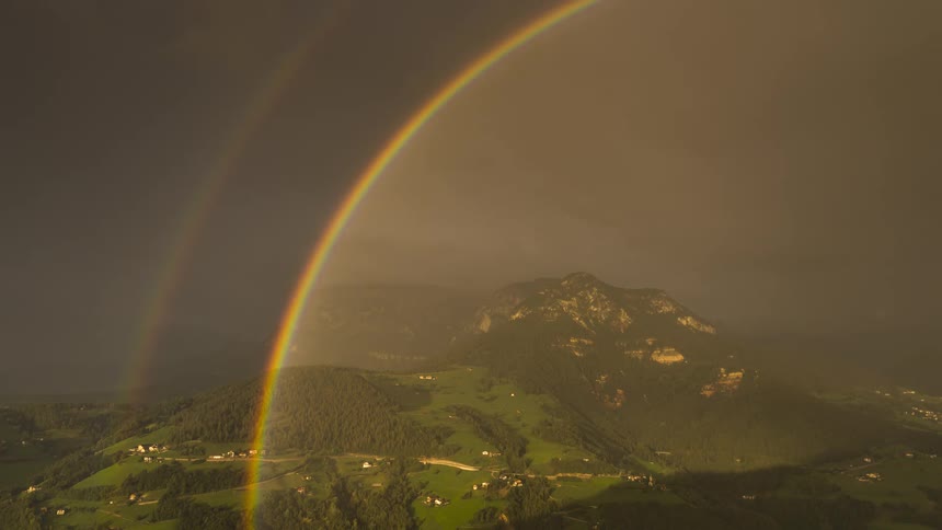 A vibrant double rainbow arches across a stormy sky over a sunlit green mountain valley