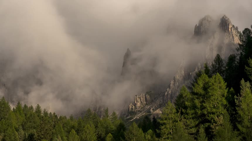 A stunning timelapse of dense fog clearing to reveal the jagged, rocky peaks of a mountain range above a lush evergreen forest