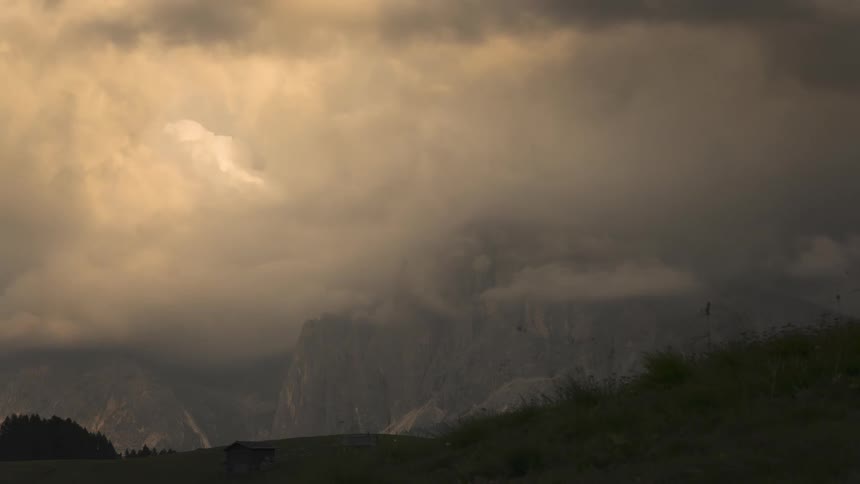 Ominous storm clouds roll over majestic mountain peaks as the sun sets, creating a dramatic and moody time-lapse scene