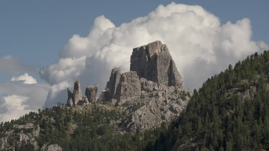 Dramatic white clouds drift behind the majestic rocky peaks of a mountain covered in a dense green forest