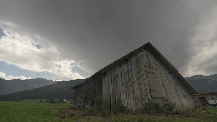 A time-lapse of dramatic storm clouds rolling over a rustic wooden barn in a mountain valley