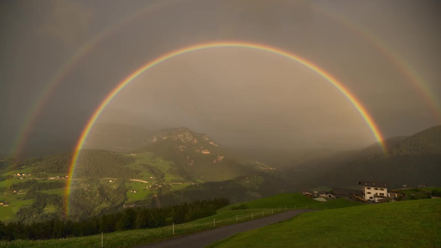 A stunning double rainbow arches over a lush green mountain valley under a dramatic, stormy sky