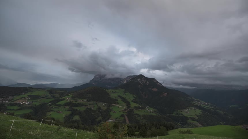 Ominous storm clouds swirl over a green alpine valley and majestic mountain range in a dramatic time-lapse