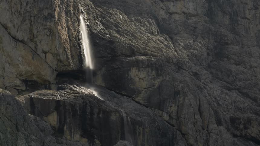 A time-lapse of a sunbeam moving across a waterfall cascading down a rugged mountain cliff
