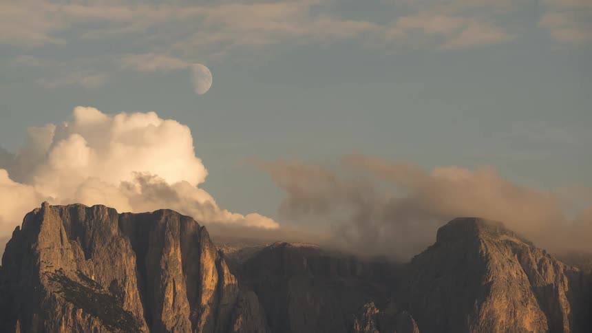 Timelapse of a waning moon in a blue sky as clouds roll over a majestic mountain range