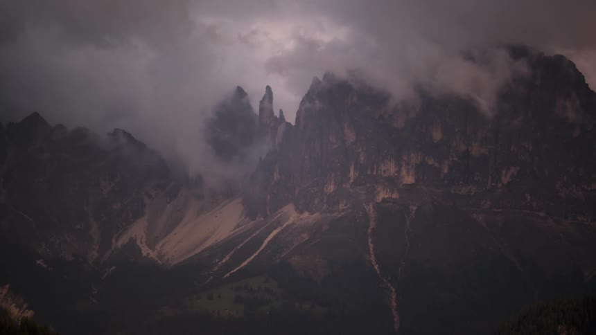 A dramatic timelapse shows clouds swirling around a majestic mountain range, revealing its jagged peaks as the sun sets