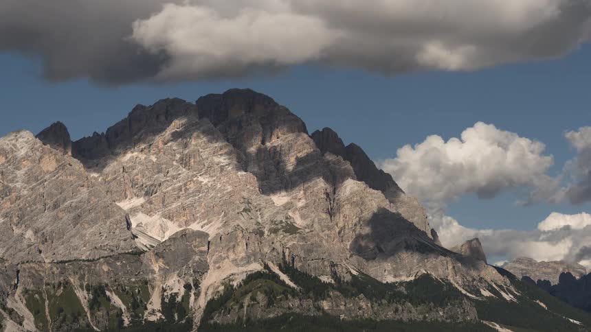 Clouds cast moving shadows across a majestic, sunlit mountain peak against a blue sky