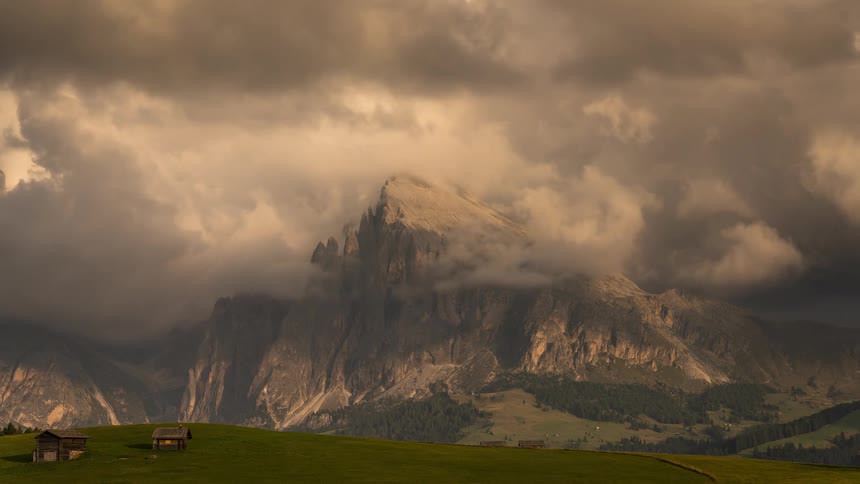 A dramatic timelapse captures clouds rolling over the jagged peaks of a mountain range in the Dolomites, with small wooden huts dotting the green meadow below