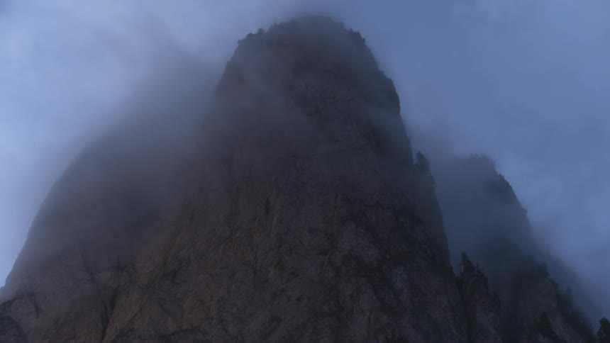 A moody time-lapse shows thick fog and clouds swirling around a majestic, rocky mountain peak in the twilight