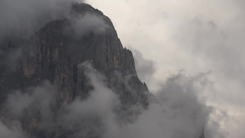 A dramatic close-up of a rugged mountain peak as thick gray clouds slowly drift by, revealing its rocky face