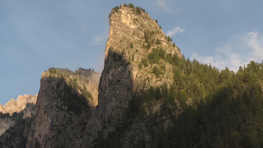 A stunning timelapse shows the setting sun casting moving shadows and golden light across a jagged, tree-covered mountain peak