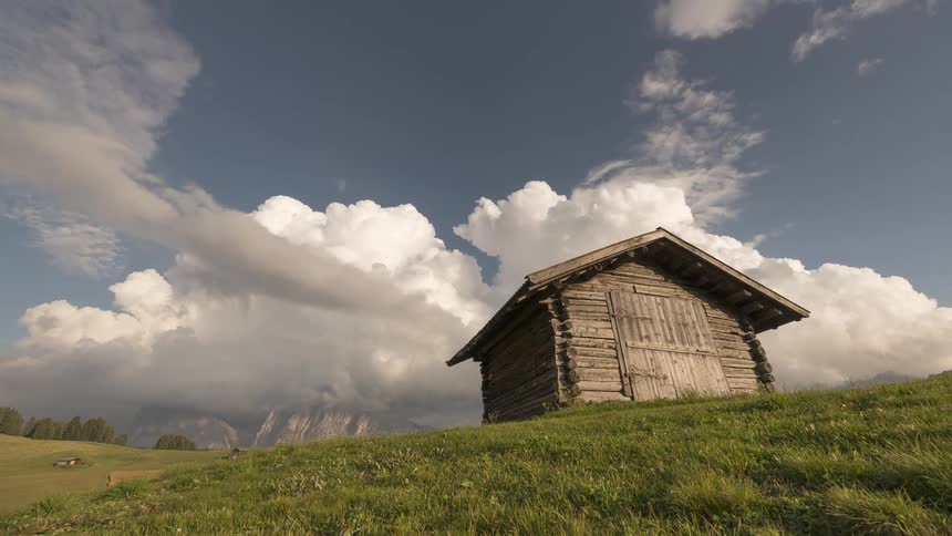 A lone wooden hut stands on a green alpine meadow as dramatic clouds drift over the mountains in a time-lapse