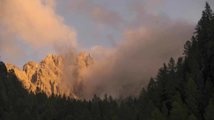 A majestic time-lapse captures clouds and fog rolling over sun-kissed mountain peaks above a dark evergreen forest