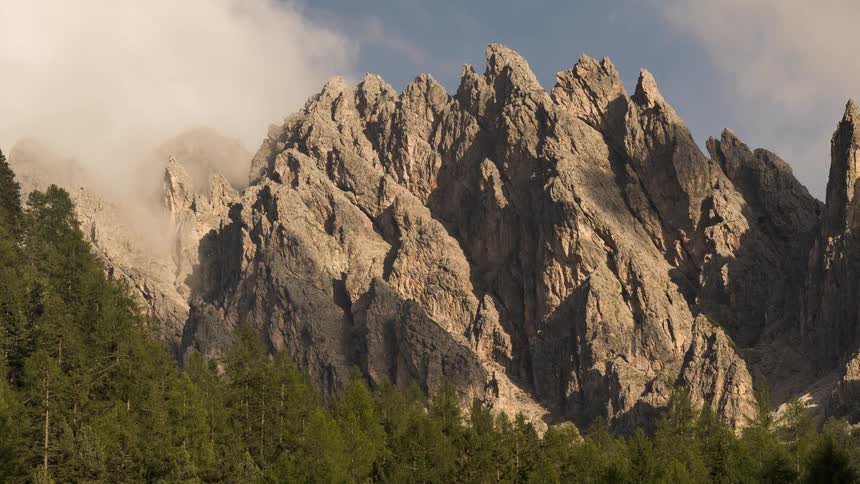 Fog and clouds dissipate in a timelapse to reveal the majestic, jagged peaks of a mountain range above a green forest