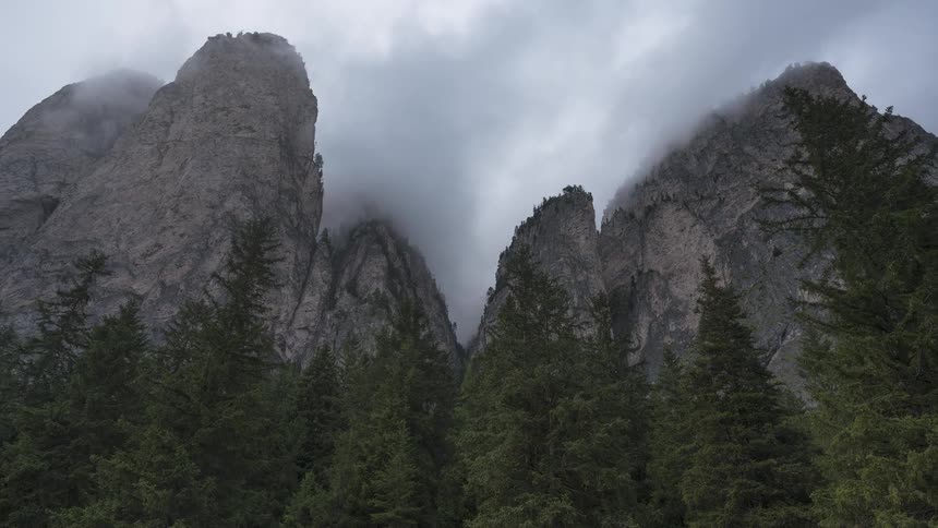 A dramatic time-lapse of thick fog and clouds swirling around towering mountain peaks above a dense pine forest
