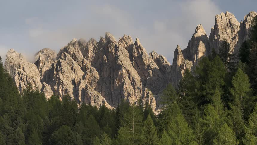 Time-lapse of clouds clearing from jagged mountain peaks that tower over a dense evergreen forest