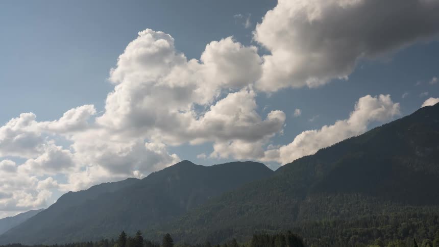 White cumulus clouds drift slowly across a blue sky above a lush, green mountain range on a peaceful day