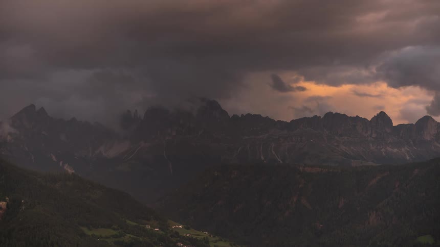 A dramatic timelapse captures dark storm clouds engulfing a golden sunset over a majestic mountain range