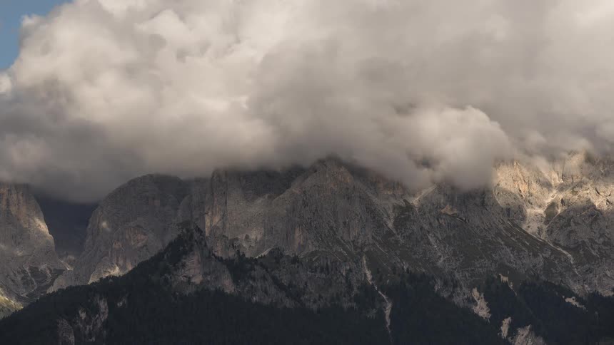 A dramatic time-lapse captures thick, moody clouds rolling over the majestic peaks of a rugged mountain range, creating a play of light and shadow