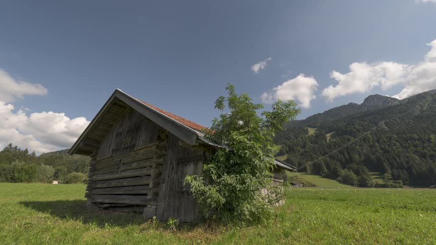 A rustic wooden shed sits peacefully in a grassy meadow with a backdrop of forested mountains under a bright blue sky